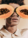 Woman holding papayas over her eyes, a key ingredient that helps ILIA Lip Wrap Hydrating Mask exfoliate lips.