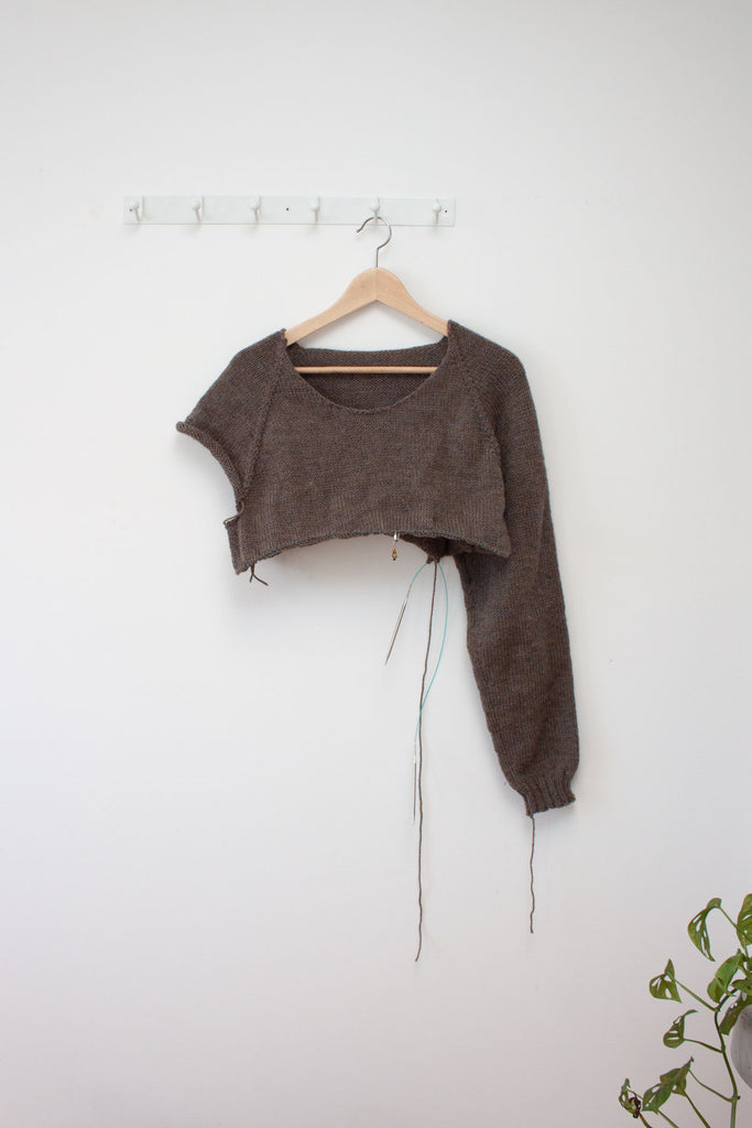 a half finished men's raglan sweater in brown hangs from a hanger against a white wall