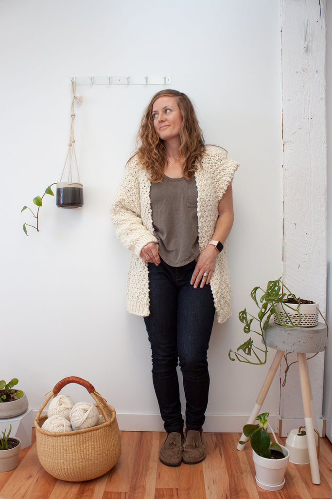 woman wearing an unfinished oversized textured handknit cardigan in a cream colour standing against a white wall