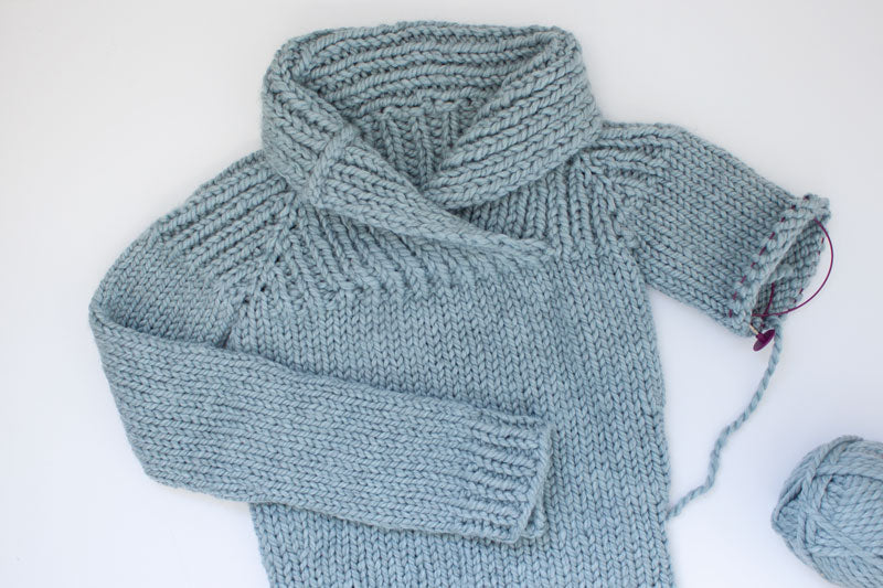 flat lay of an unfinished bulky hand knit sweater with shawl collar in grey blue