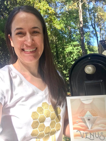 Astronomer Misty Bentz smiles in front of a mailbox holding a NASA JPL Venus Travel postcard and wearing a white v-neck JWST t-shirt