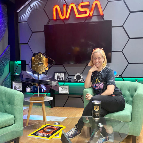 Emily sitting on a green arm chair on a talk-show-like set with lots of nerdy props and the NASA worm logo in the background.