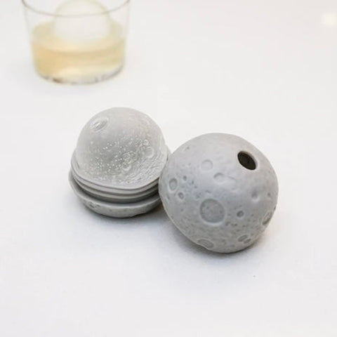 Moon Ice Ball Mold shown open filled with an ice ball, with a whickey glassin the background, on a white surface