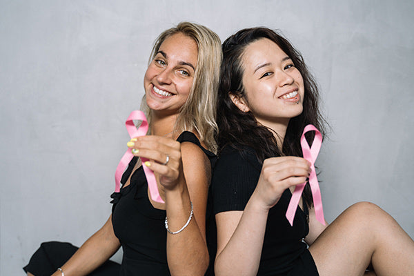 the benefits of white fir essential oil two woman sitting back to back wearing black smiling holding pink ribbons for cancer awareness light gray background