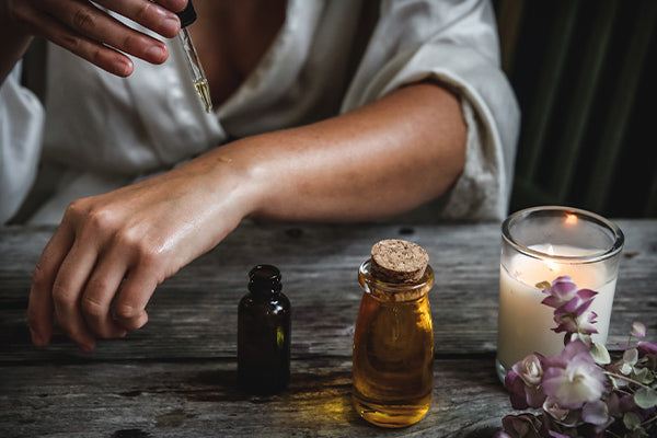 the benefits and uses of black spruce essential oil person woman in white robe applying black spruce oil topically on arm oil flowers and candle sitting on wood table