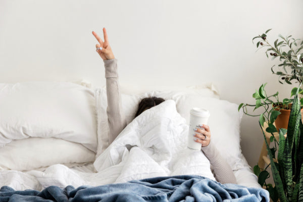 the beginners guide to improving well being person laying on bed with sheet over face holding up coffee peace sign