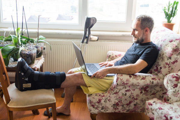 tendonitis vs tendinosis what is the difference man sitting on flower print arm chair with foot elevated in a boot for tendonitis working on a laptop crutches beside him