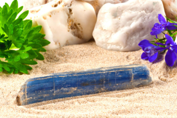the healing properties of blue kyanite bar of raw blue kyanite sitting in the sand with rocks leaves in background