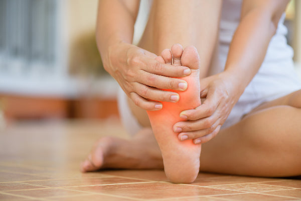 what is metatarsalgia metatarsalgia symptoms causes natural remedies woman wearing white sitting on tile floor holding the ball of her foot in pain red pain orb