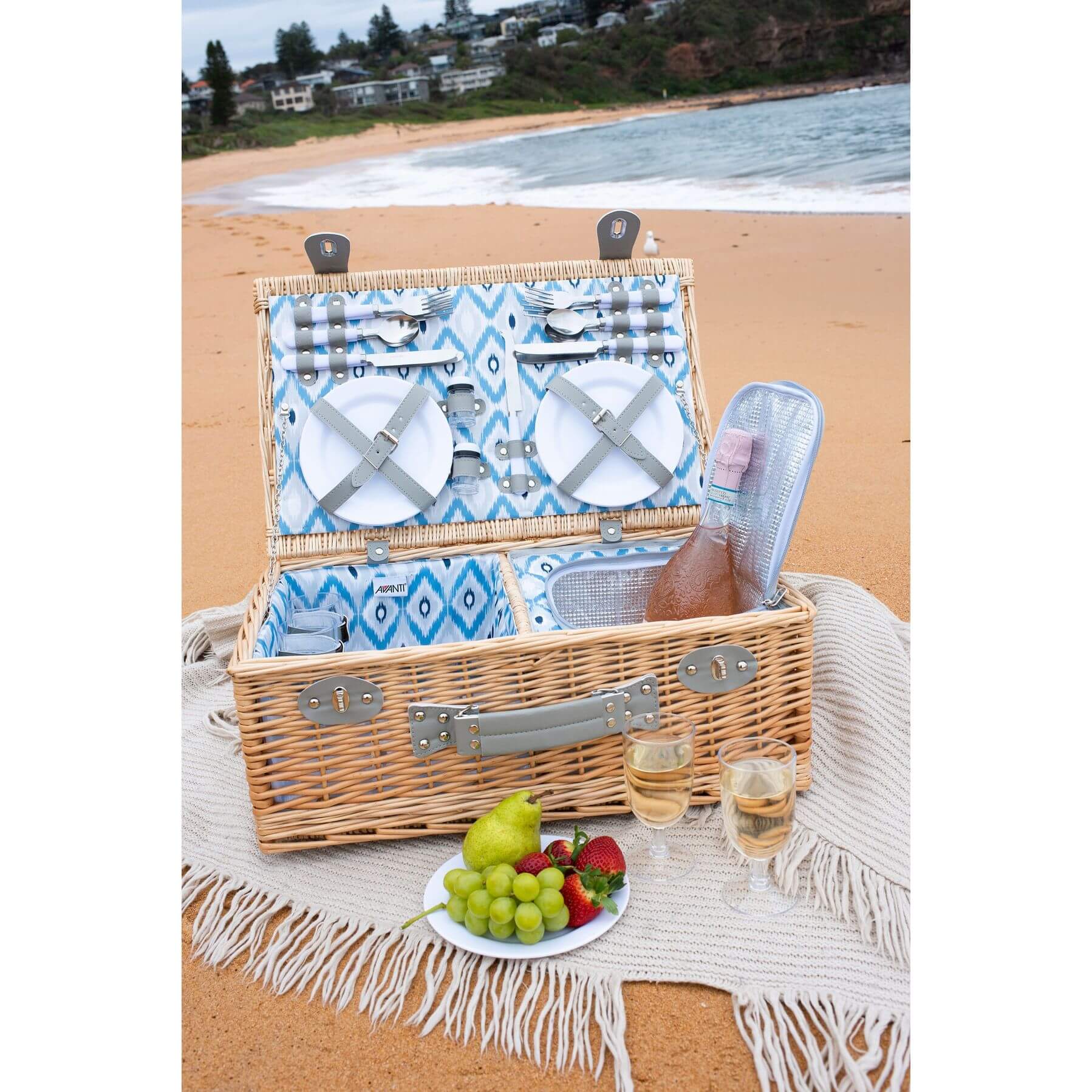 A wicker insulated picnic basket on a beach with food, wine and a picnic rug