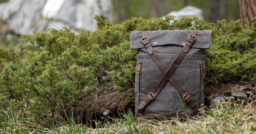 vintage rugged heavy duty canvas and brown full grain leather backpack with padded adjustable shoulder straps laying on a grass floor in front of a bush