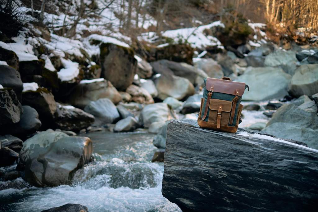 sac à dos vintage pour homme de randonnée ou camping en toile cirée et cuir de vache café posé sur un rocher près d'une rivière