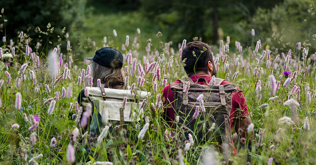 deux randonneurs portant des sacs à dos vintage en toile canvas et cuir pleine fleur marron dans un champ de fleurs sauvages pendant leur voyages