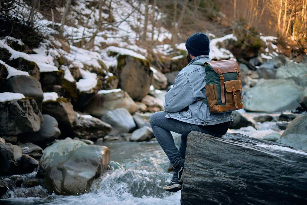 randonneur assis sur un rocher près d'une rivière en hiver portant un grand sac à dos vintage en cuir pleine fleur brun et toile cirée émeraude