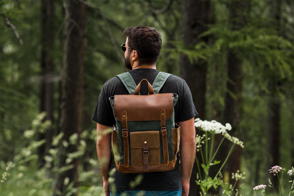 homme brun de dos dans une fôret portant un sac à dos baroudeur vintage en toile cirée vert émeraude et cuir marron