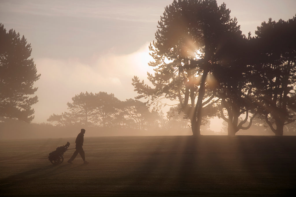 Side vie of man walking past trees across golf course at twilight, pulling golf trolley.