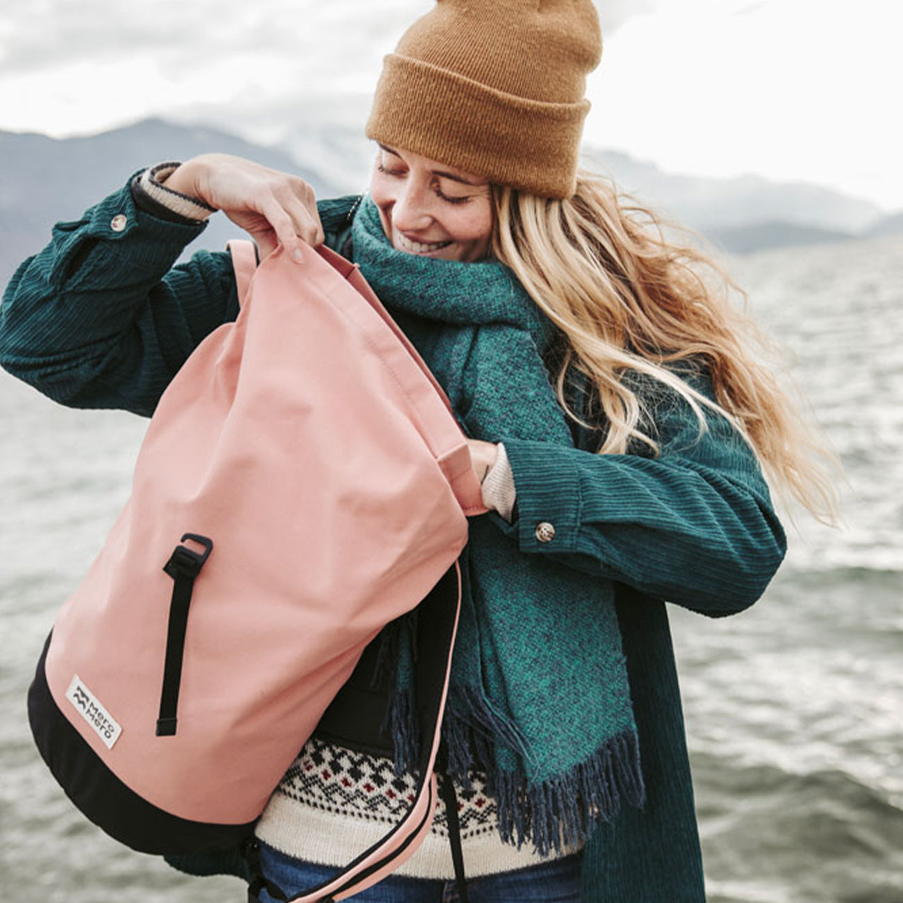woman in front of a lake wearing a pink roll top eco friendly backpack from mero mero