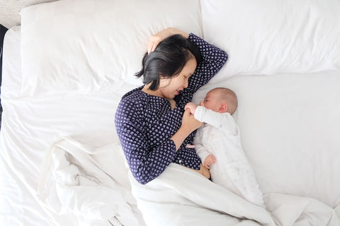 Woman lying in a big bed with white sheets and playing with her baby