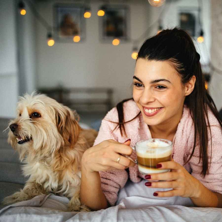 Young woman lying on her bed drinking coffee and giggling with her small dog as they celebrate National Best Friends Day!