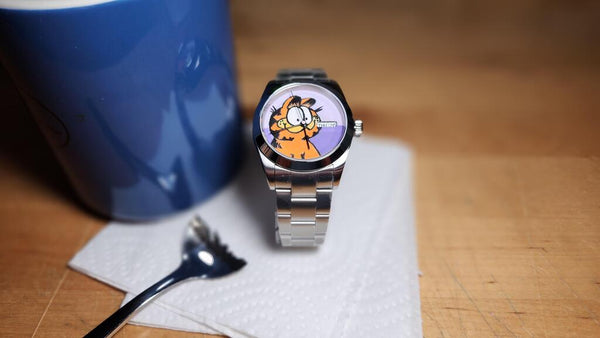 minimalist dress watch with a garfield cartoon cat dial next to a blue mug