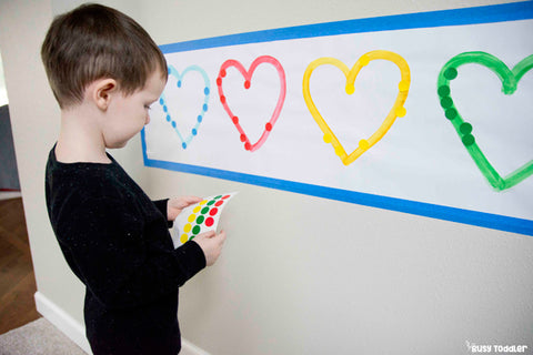 A toddler placing stickers on a piece of paper with four hearts drawn on it