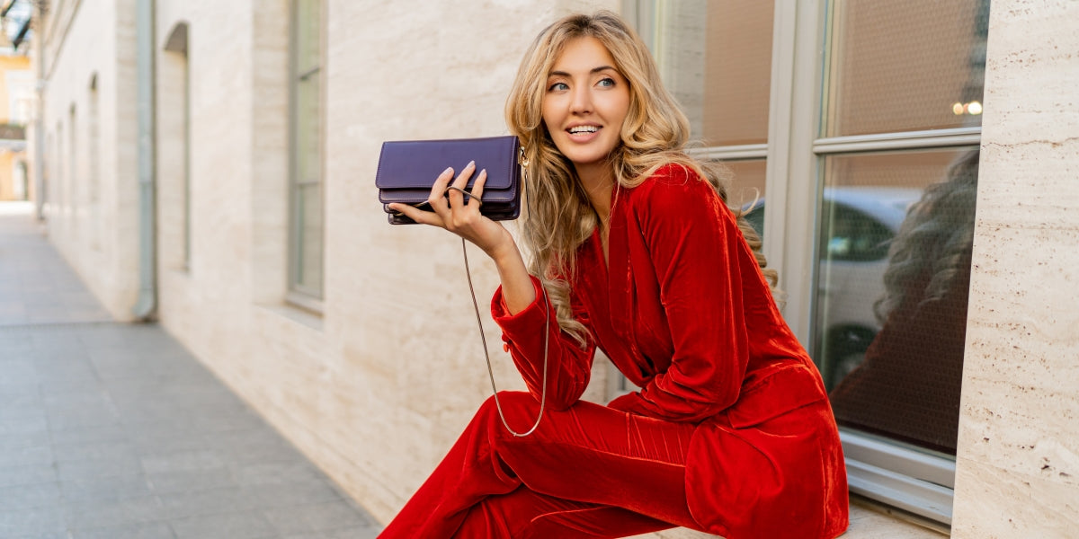 Beautiful smiling woman in elegant red velvet suit holding purse and posin outdoor in old european city. Blond wavy hairs, perfet skin, full lips.
