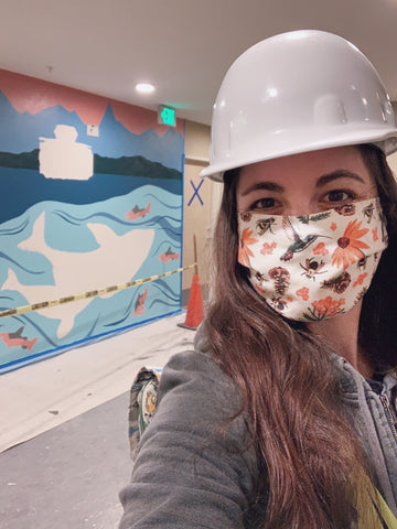 Woman in a hard hat and mask stands in front of a mural that is in progress. The mural is of a Washington State Ferry with the Olympic mountains in the background and orcas under in the water.