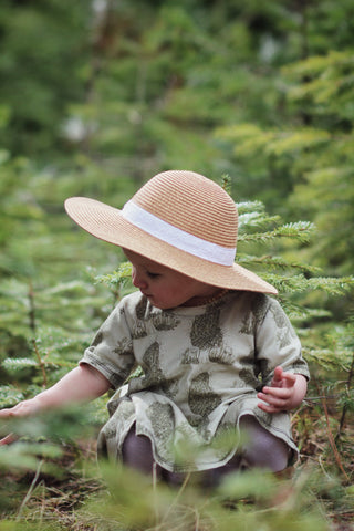 A little girl sits in the grass with a morel dress on. She is picking some foliage with a pine tree in the background.