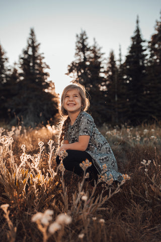 Little girl in a grey raccoon dress sits in a flowery field with trees behind her smiling at the sky.