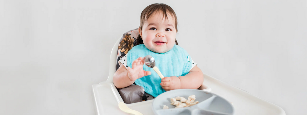 toddler sitting in a high chair with food and holding a spoon