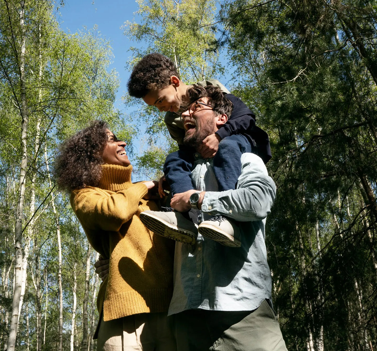 Smiling mum looking up at a young boy on his father’s shoulders, on a sunny walk among the birch trees