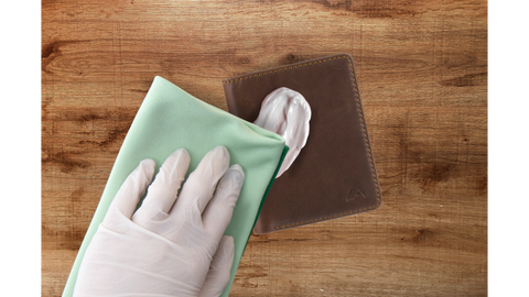 A hand wearing a plastic glove wipes a green cloth over white cleaning product into a brown wallet