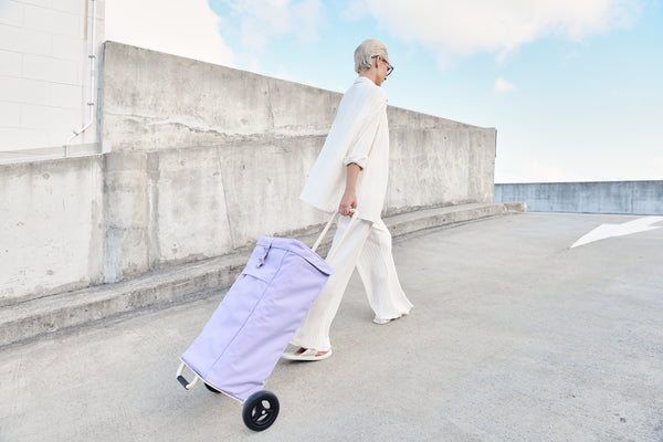 A women wearing a beige matching pant suit pulling a lilac Joyrolla Shopping Cart in a concrete shopping centre car park