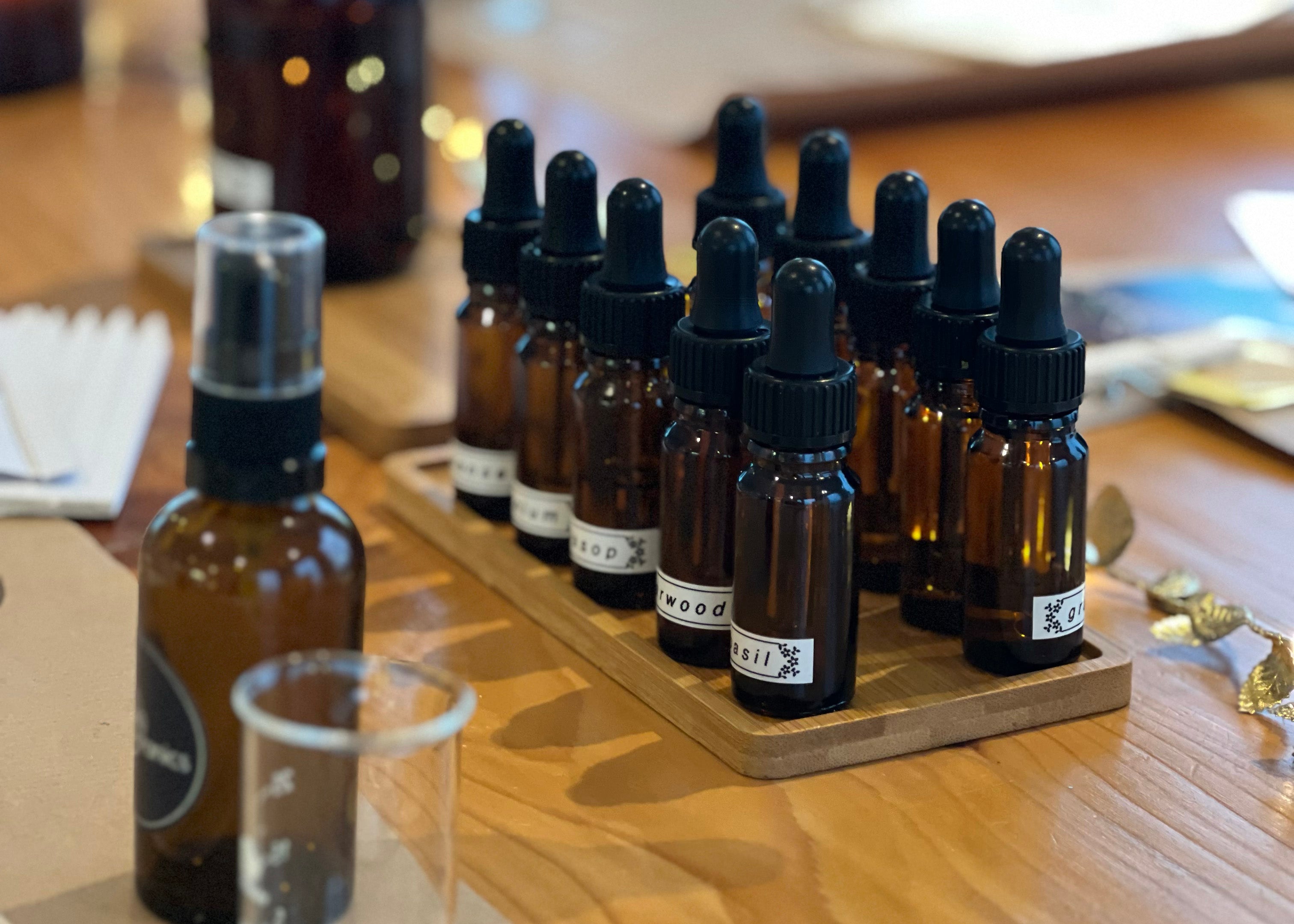 A collection of amber glass bottles with droppers, labeled with various scents such as 'cedarwood' and 'basil', are neatly arranged on a wooden tray. In the foreground, a brown glass spray bottle and a clear measuring cup are visible, suggesting a workshop setting for creating custom scents or aromatherapy blends