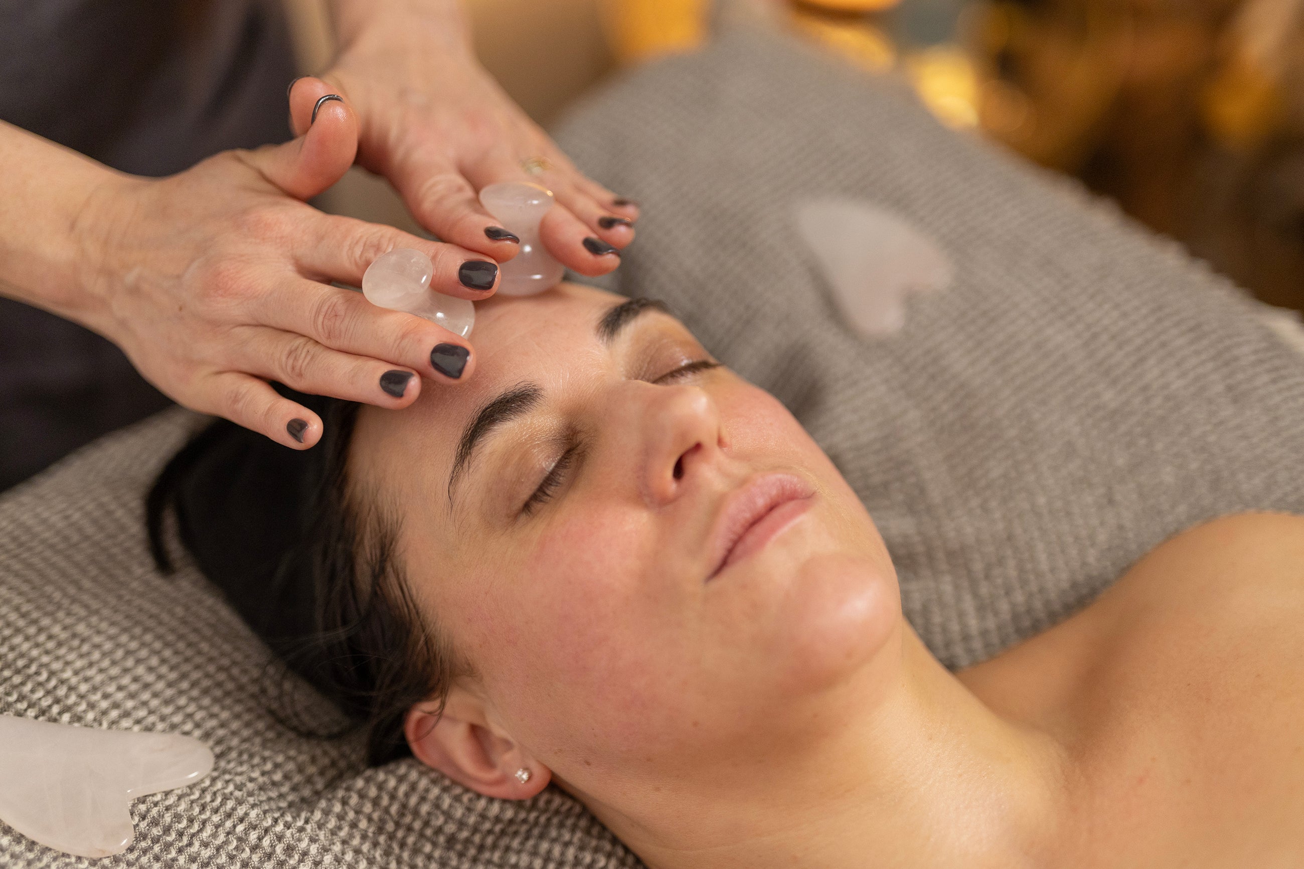 A person receiving a crystal facial massage, with eyes closed and clear quartz stones placed on their forehead by a practitioner with polished nails, in a serene spa environment.