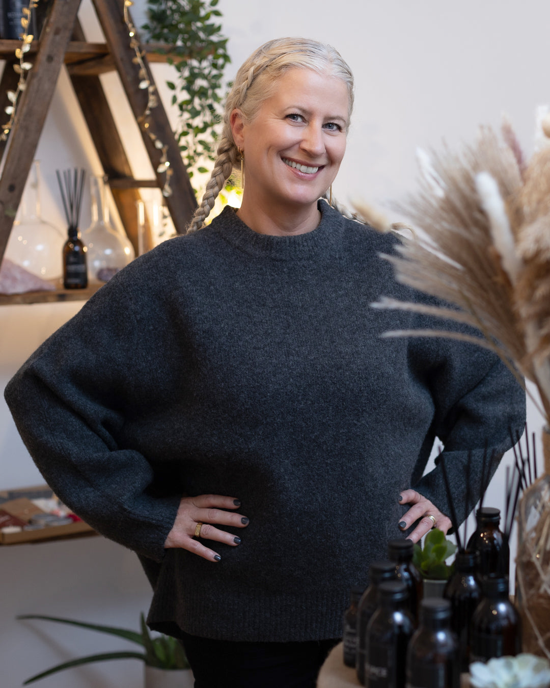 A woman with a warm smile stands in a cosy, naturally lit room. Her hair is styled in elegant, tight braids wrapped around her head, highlighting her silver strands. She's wearing a comfortable, oversized charcoal sweater and black trousers, with her hands placed confidently on her hips. In the background, there's an assortment of stylish decor including pampas grass, delicate string lights, glass vases, and an array of dark bottles, possibly containing artisanal products, which create an inviting and tranquil atmosphere