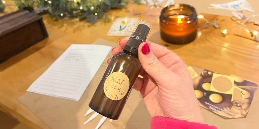 A close-up view of a hand holding a brown glass spray bottle with a handwritten label "Wild Love 22.01.24", suggesting a personalized aromatherapy or natural wellness product created at a workshop. The hand is adorned with red nail polish, adding a pop of color to the image. In the background, there's a workshop table with papers, wooden sticks, fairy lights, and a large amber candle, all contributing to a warm and engaging DIY crafting experience.
