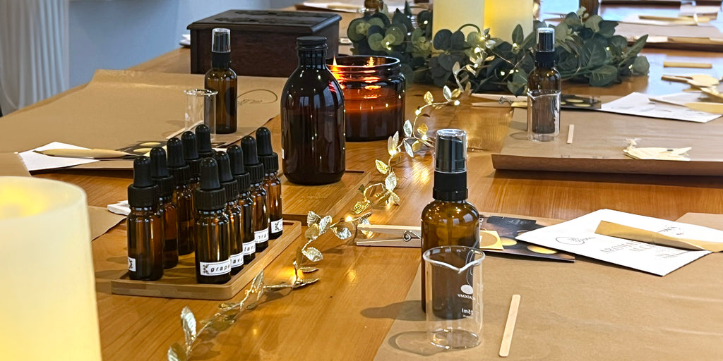 A well-organized workshop table displaying an array of amber glass bottles in various sizes, some with dropper caps, amidst a creative setting. A measuring glass, wooden spatulas, and informative cards are laid out for participants. The ambiance is enhanced by a glowing candle and fairy lights woven through eucalyptus branches, indicating a focus on natural wellness and beauty products. The scene conveys a blend of education and relaxation, typical of a wellness retreat workshop.