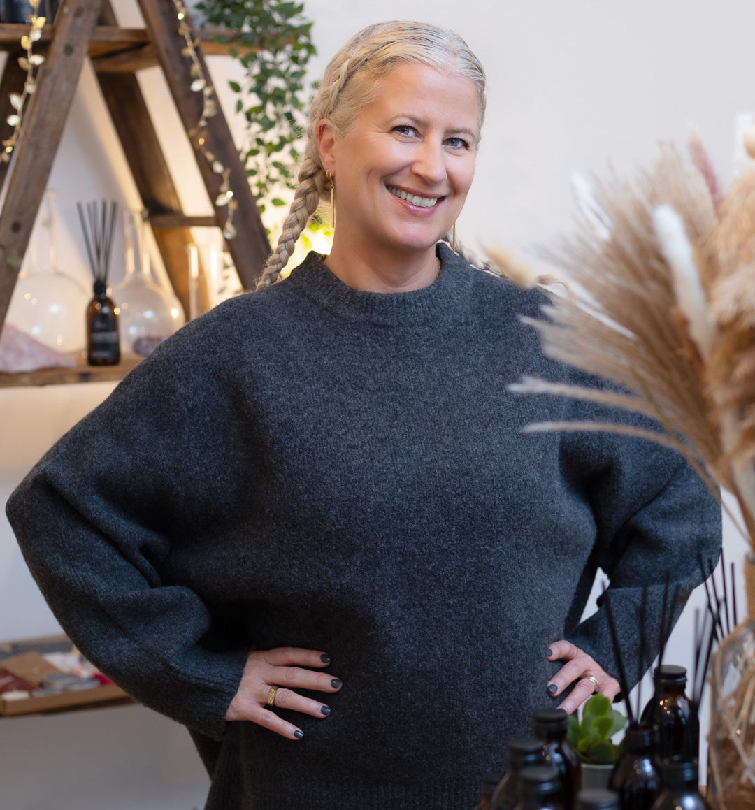 A confident woman with a beaming smile and silver braided hair poses in a cozy charcoal grey sweater. She stands in a workshop space adorned with dried pampas grass and a wooden ladder festooned with fairy lights. In the background, an assortment of amber glass bottles and aroma diffusers on a shelf contribute to the warm, inviting ambiance of the natural skincare workshop