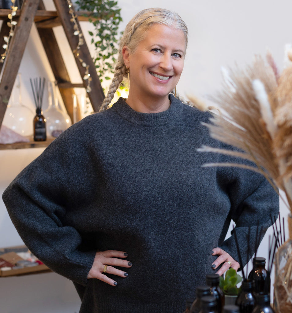 Confident woman with braided hair smiling in a cozy wellness shop, surrounded by artisanal aromatherapy products and natural decor, embodying a holistic and inviting atmosphere.