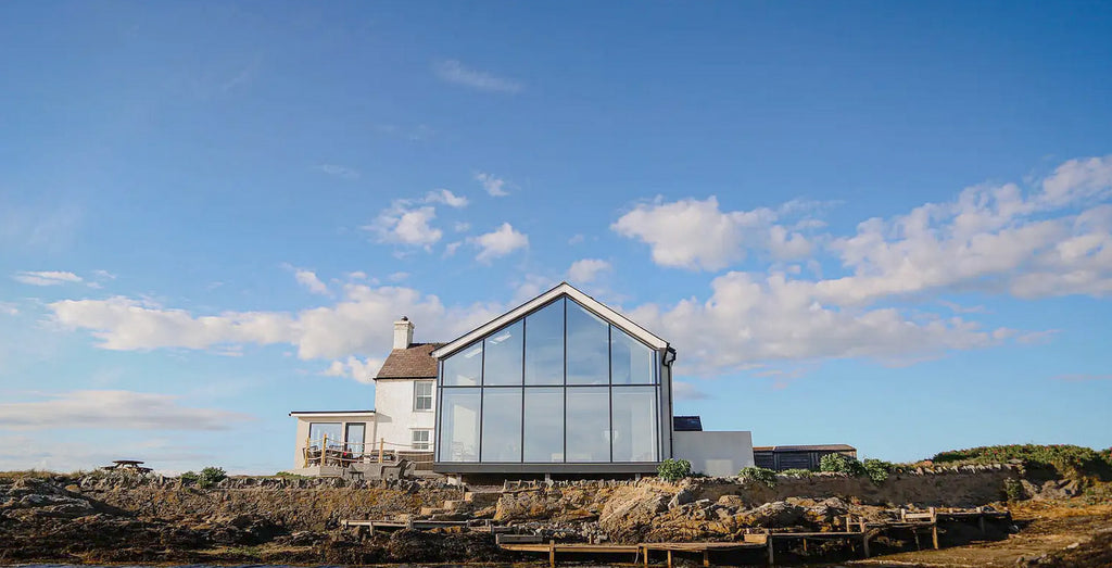 A modern coastal retreat house in Wales, featuring a large glass facade extension alongside a traditional white cottage structure. The house is perched on a rugged rocky landscape with wooden walkways, under a vast blue sky with scattered clouds. The serene setting and contemporary architectural design suggest a blend of comfort and style, ideal for a wellness workshop or retreat