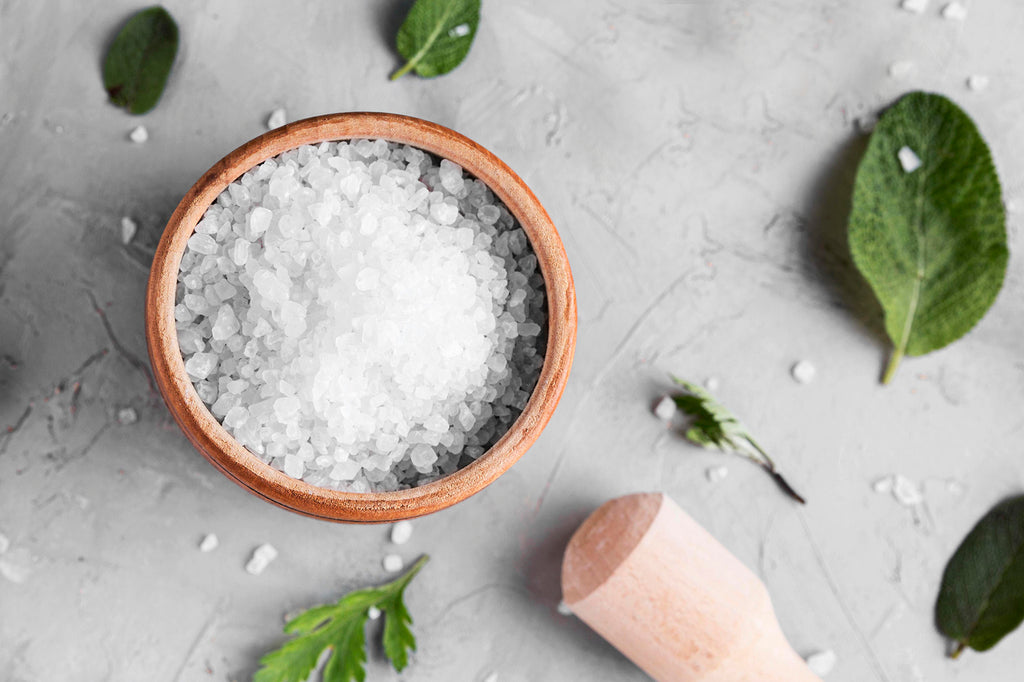 salt in wooden bowl on white surface with scattered herb leaves