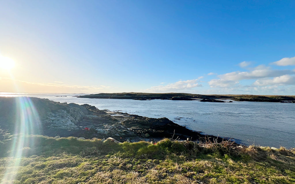 A serene landscape view of the rugged Welsh coastline with the sun hanging low in the sky, casting a bright glare and reflecting off the calm sea. The coastline is dotted with rocky outcrops and sparse vegetation, under a blue sky with light cloud cover. The scene conveys a sense of tranquility and natural beauty, typical of a coastal retreat in Wales