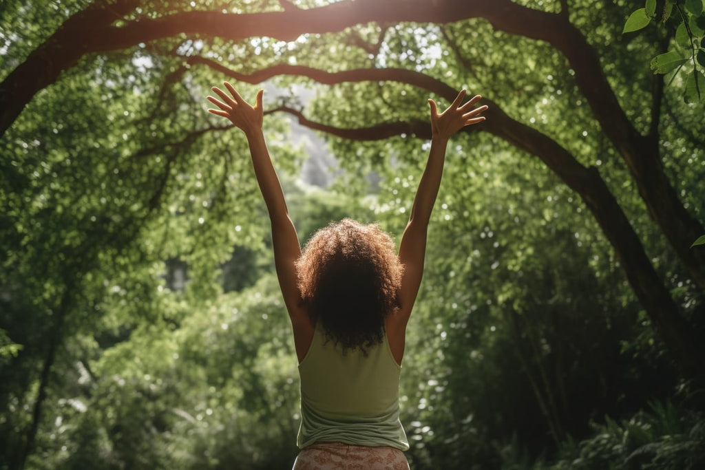 A person in a natural outdoor setting, surrounded by plants and trees, stretches their arms overhead in a peaceful yoga pose.