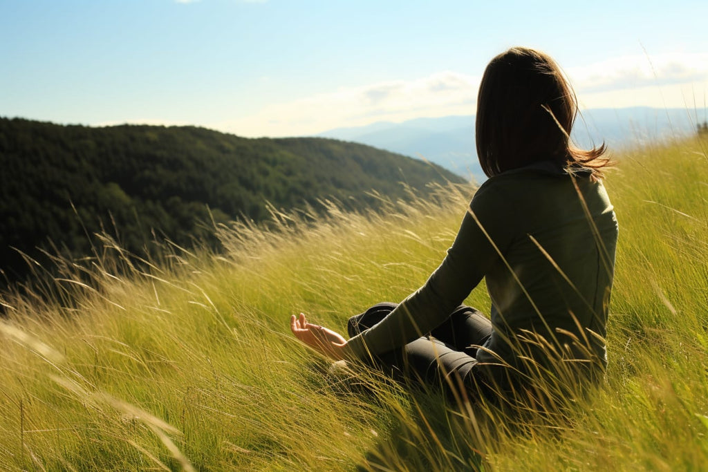 A person sitting cross-legged on a grassy hillside with hands placed firmly on their lower back, feeling the warmth of the sun and the breeze of a summer day.