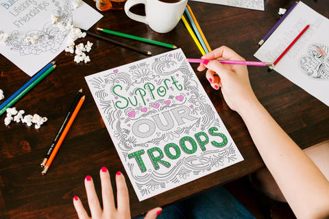 coloring page that reads "support our troops" partially colored in pink and green, sitting on a tabletop that is also holding colored pencils, popcorn, a cup of coffee and a variety of coloring pages, with a woman's hands holding a pink colored pencil