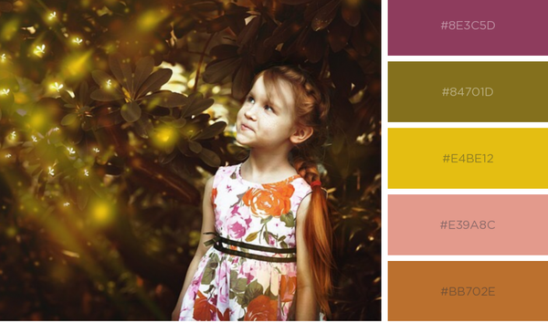 Young girl with red braided hair, wearing dress with coral and pick flowers, standing in front of tall shrub with dots of sunlight shining through.