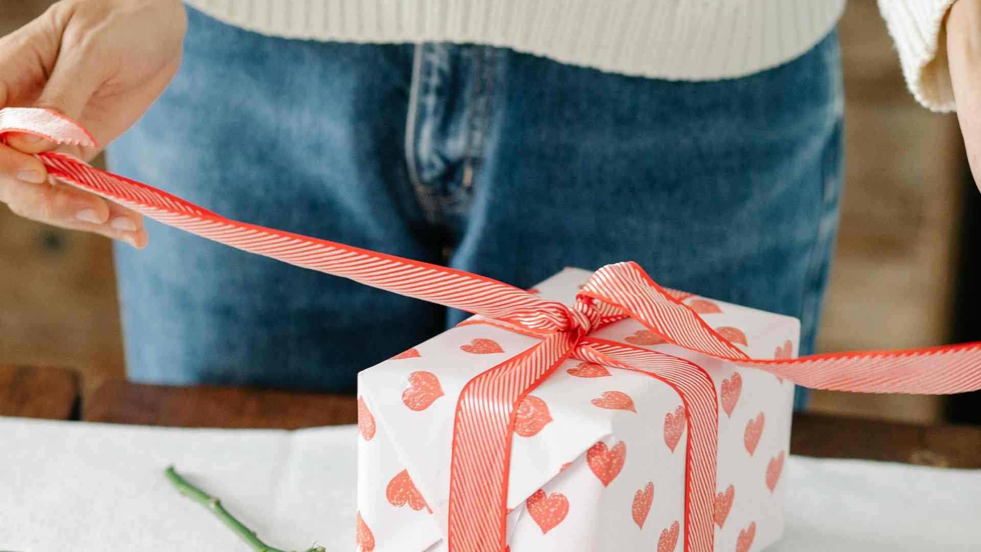 Rankos, rišančios raudonai baltai dryžuotą juostelę ant baltos dovanų dėžutės su raudonomis širdelėmis, pastatytos ant medinio paviršiaus.