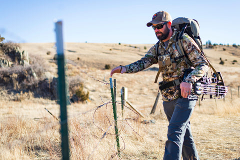 Bow hunter wearing camo with a bow sling crossing a fence in Eastern Montana