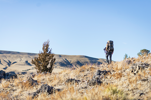 Hunter overlooking a bluff in Montana on a blue sky day wearing a Mystery Ranch backpack with rifle attached to his pack using a hands-free rifle sling